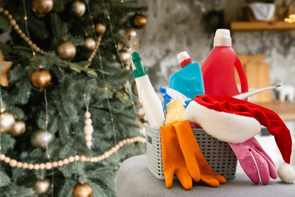 A festive living room with a decorated Christmas tree in the background and a basket of cleaning supplies on the couch, preparing the home for the holiday season.